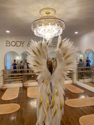 Glam barre studio with oversized white-and-gold angel wings under a crystal chandelier, mirrored wall, ballet barres and workout mats
