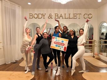 Cheerful group of dancers posing at a boutique ballet studio barre, arms raised in ballet poses in front of mirrors, gold wall lettering and a colorful poster.