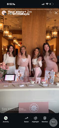 Four smiling women in pink and white activewear waving behind a pastel-pink wellness pop-up table with brochures, QR codes, flowers and a tablet in a warm, lantern-lit event space.