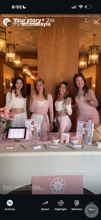 Four smiling women in pink and white activewear waving behind a pastel-pink wellness pop-up table with brochures, QR codes, flowers and a tablet in a warm, lantern-lit event space.