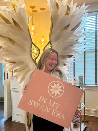 Smiling person posing with oversized white-and-gold feather wings, holding a pink sign that reads "IN MY SWAN ERA" in a warm-lit indoor setting with globe pendant lights and window blinds.