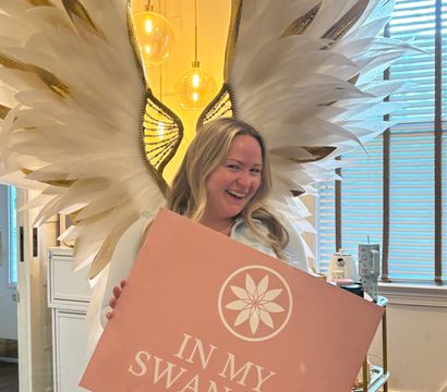 Smiling person posing with oversized white-and-gold feather wings, holding a pink sign that reads "IN MY SWAN ERA" in a warm-lit indoor setting with globe pendant lights and window blinds.