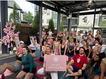 Cheerful outdoor group yoga/fitness class on a covered urban patio at an apartment complex, participants smiling on mats around a pink sign reading IN MY SWAN ERA and decorative blossoms