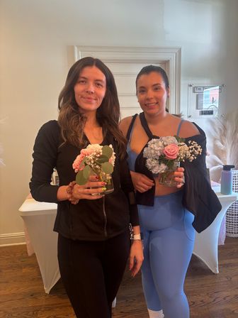 Two smiling women in casual activewear holding small glass vases of pink roses, hydrangeas and eucalyptus indoors beside pampas grass — bright studio portrait.