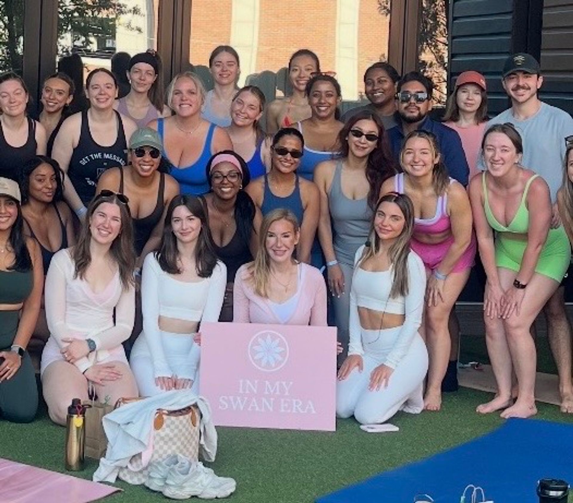 Large diverse group in colorful activewear posing on an outdoor lawn in front of patio doors, holding a pink sign that reads "IN MY SWAN ERA" at a playful group fitness/yoga event