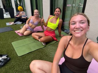 Three women in colorful activewear smiling for a selfie during an outdoor patio yoga session on artificial turf by glass doors, sitting on mats with one giving a thumbs-up.