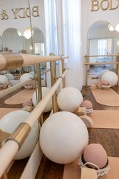 Light-filled boutique barre studio interior with gold and wood barres, arched mirrors and sheer curtains, neutral cork mats, large white exercise balls and pink Pilates balls neatly arranged.