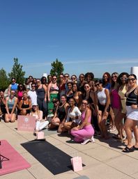 Large diverse group in activewear posing on a sunny rooftop terrace for an outdoor yoga/fitness event with yoga mats, pink gift bags and trees under a clear blue sky.
