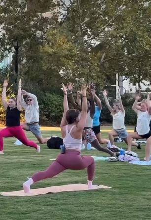 Outdoor group yoga class in a park, people on mats practicing crescent lunges with arms raised on a grassy lawn under trees, casual fitness gear and water bottles visible.