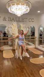 Joyful woman in pale blue workout set strikes a ballerina pose on one leg wearing oversized white-and-gold feather wings in a mirrored ballet studio beneath a crystal chandelier, with hardwood floors and barres visible.