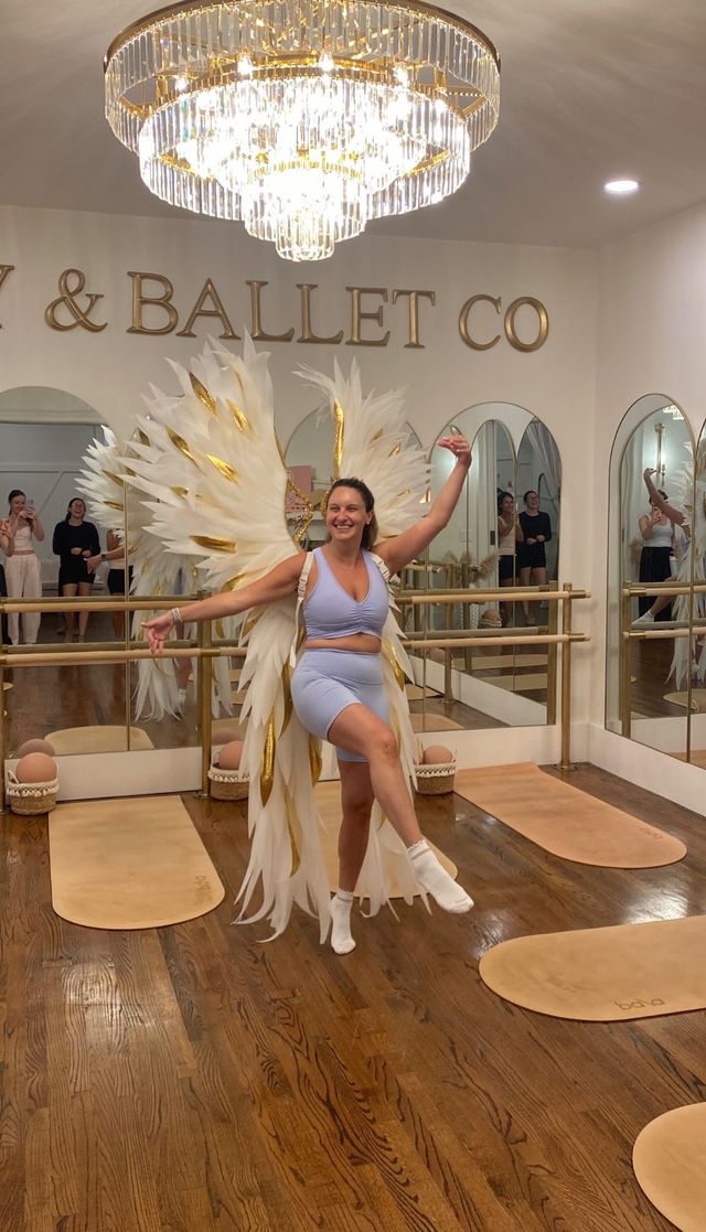 Joyful woman in pale blue workout set strikes a ballerina pose on one leg wearing oversized white-and-gold feather wings in a mirrored ballet studio beneath a crystal chandelier, with hardwood floors and barres visible.