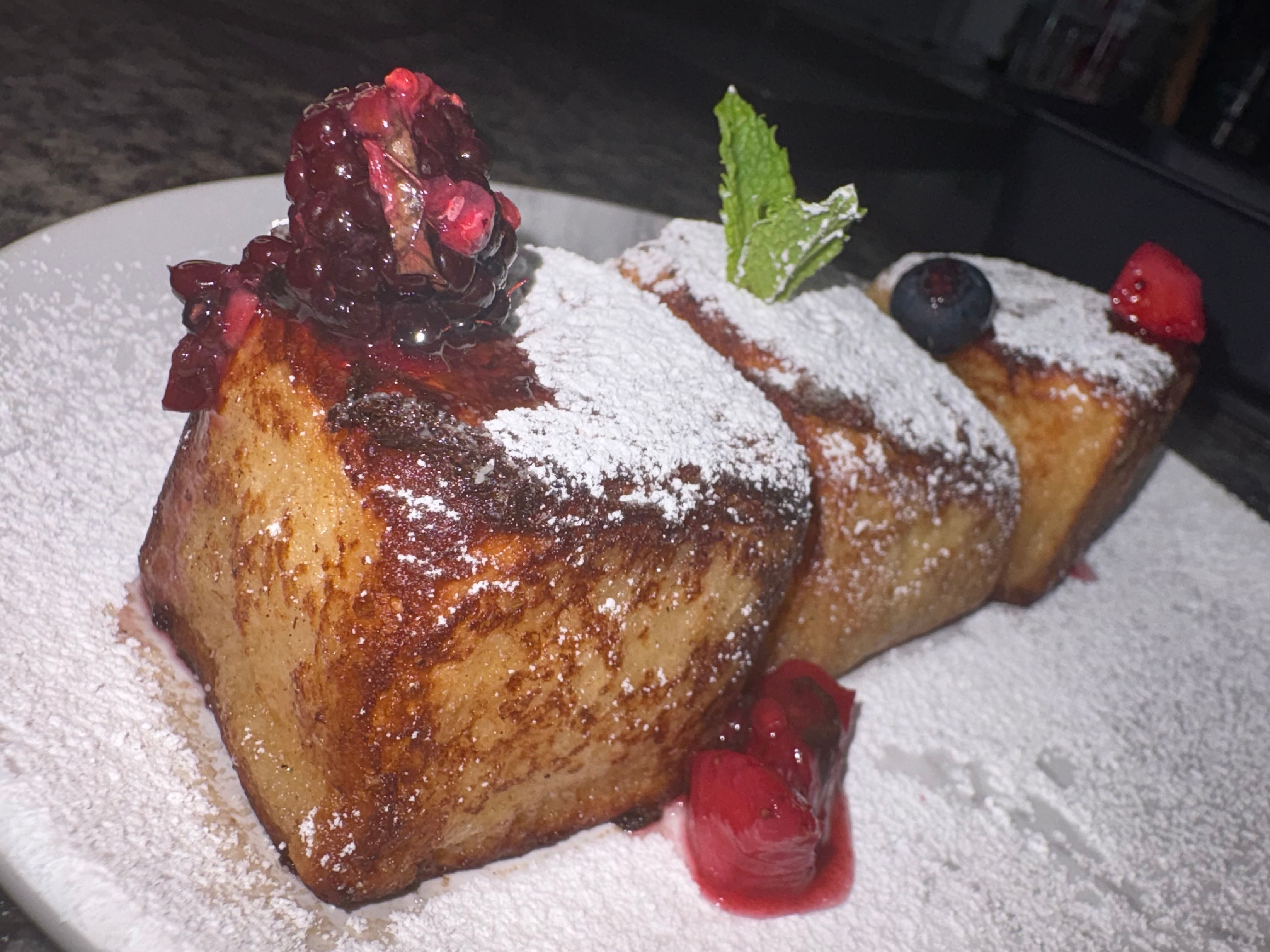 Close-up of three thick cinnamon-browned brioche French toast cubes dusted with powdered sugar, topped with blackberry compote, blueberry, mint leaf and cherries on a white plate — brunch dessert