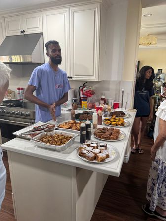 Man serving a homemade brunch spread on a white kitchen island — trays of powdered pastries, French toast, breakfast potatoes, grilled items and squeeze bottles of sauces, with white cabinets, stainless range and guests nearby.