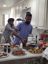 Two people preparing a home kitchen brunch spread on a kitchen island — trays of homemade pastries, a skillet of savory bites, and squeeze bottles of sauces for a casual breakfast buffet.
