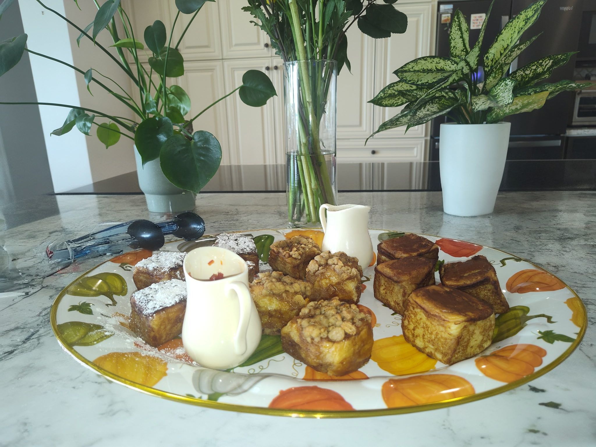 Assorted bite-sized French toast cubes and streusel-topped pastries on a decorative fall-themed plate with two mini cream pitchers, set on a marble kitchen island with potted plants and a vase of flowers in the background.