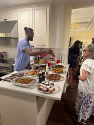 Home kitchen brunch buffet on a white island: trays of roasted potatoes, French toast, powdered-sugar cinnamon rolls, muffins and bottles of syrup and sauces; a man in a blue shirt pours sauce while two guests wait nearby.
