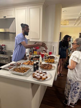 Home kitchen brunch buffet on a white island: trays of roasted potatoes, French toast, powdered-sugar cinnamon rolls, muffins and bottles of syrup and sauces; a man in a blue shirt pours sauce while two guests wait nearby.