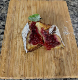 Toasted brioche slice oozing bright cranberry jam, dusted with powdered sugar and a mint leaf on a bamboo cutting board over granite countertop