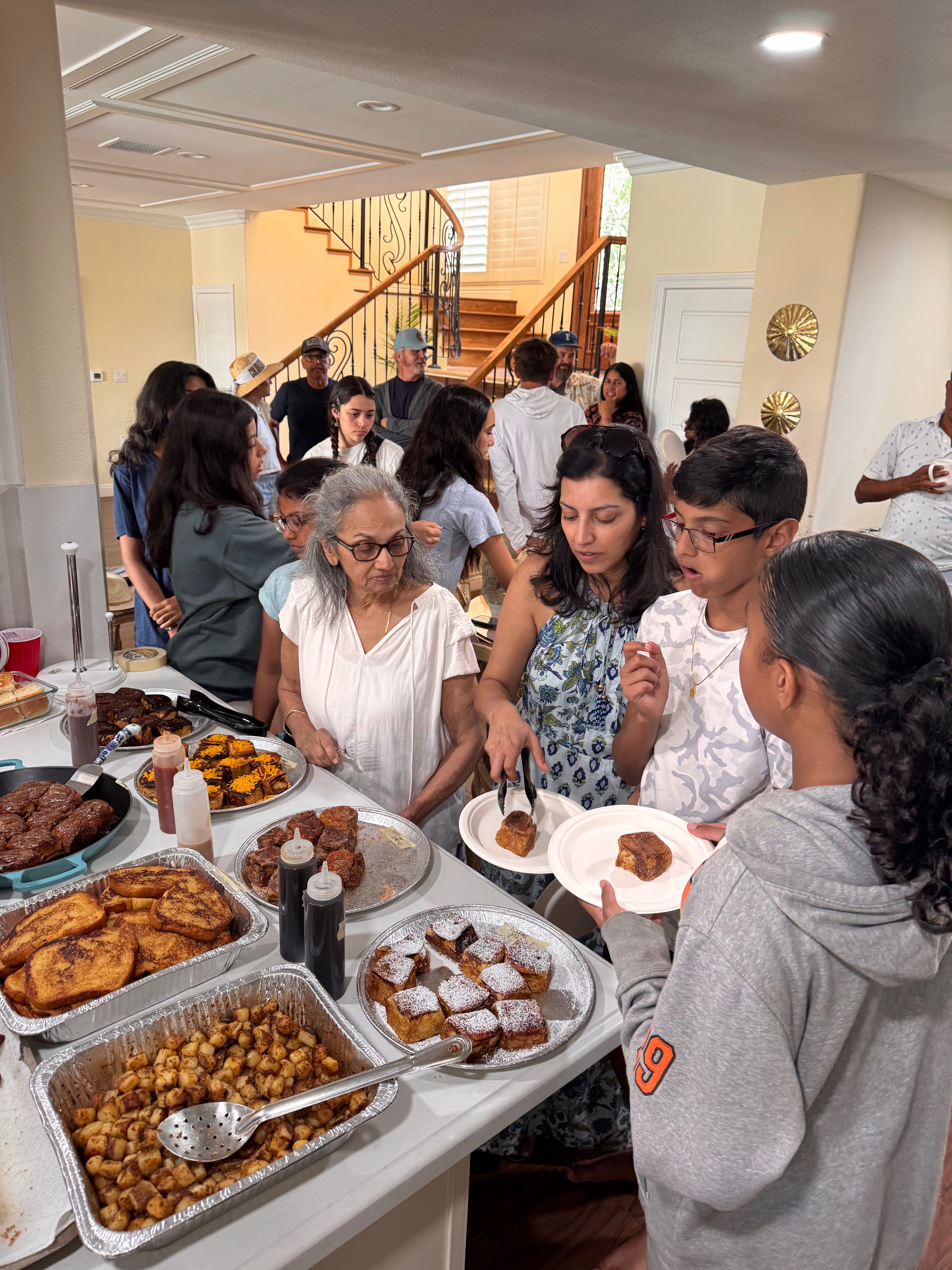 Busy home kitchen brunch buffet with a multi-generational family serving French toast, powdered pastries and breakfast potatoes near a wooden staircase.