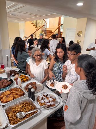 Busy home kitchen brunch buffet with a multi-generational family serving French toast, powdered pastries and breakfast potatoes near a wooden staircase.