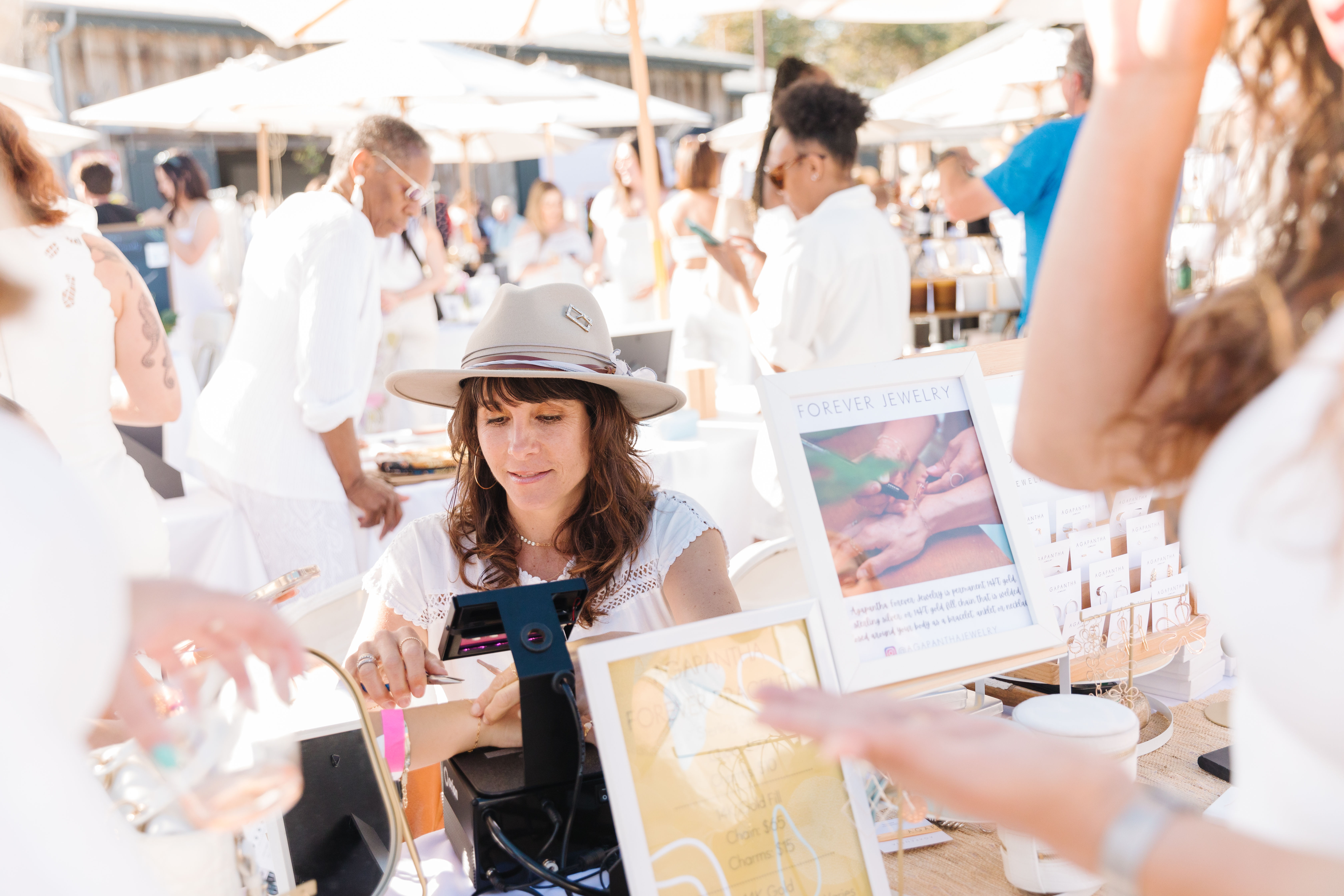 Vendor in a tan hat demonstrating artisan jewelry at a sunny outdoor market pop-up, customers browsing booths under white umbrellas