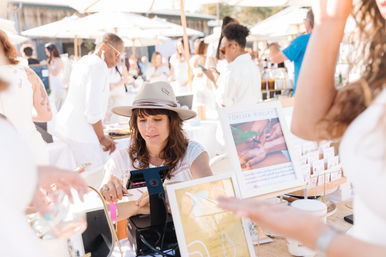 Vendor in a tan hat demonstrating artisan jewelry at a sunny outdoor market pop-up, customers browsing booths under white umbrellas