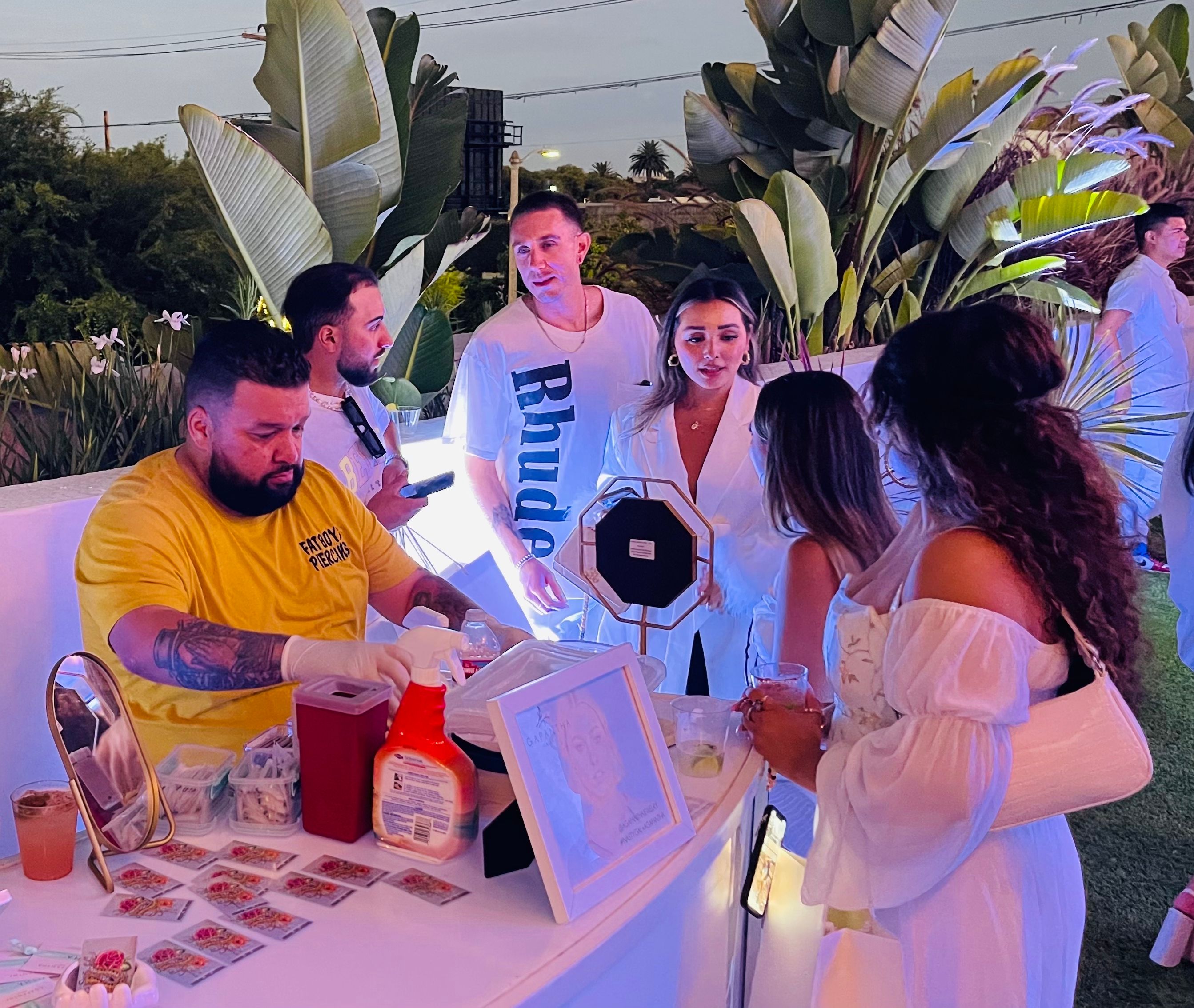 Evening outdoor party guests in white gather around an illuminated pop-up vendor table with mirrors, framed art and supplies; a tattooed vendor in a yellow shirt serves attendees beneath tropical plants and purple-blue uplighting.