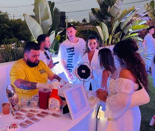 Evening outdoor party guests in white gather around an illuminated pop-up vendor table with mirrors, framed art and supplies; a tattooed vendor in a yellow shirt serves attendees beneath tropical plants and purple-blue uplighting.