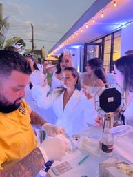 Evening backyard party with friends in white outfits raising cocktails under string lights while a gloved bartender in a yellow shirt mixes drinks at a modern patio bar.
