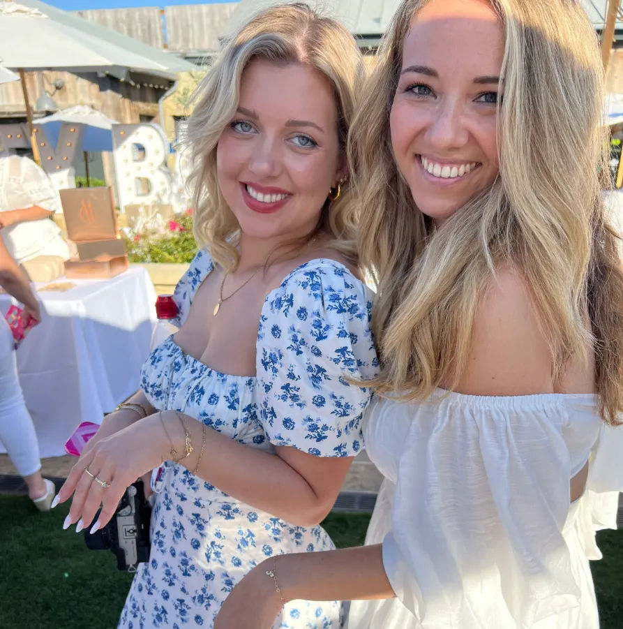 Two smiling women posing at a sunlit outdoor summer party — one in a blue floral dress and the other in an off-shoulder white dress, with tables and marquee letters in the backyard background.
