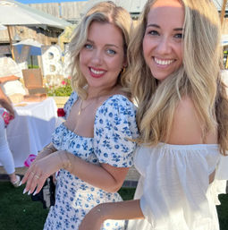Two smiling women posing at a sunlit outdoor summer party — one in a blue floral dress and the other in an off-shoulder white dress, with tables and marquee letters in the backyard background.