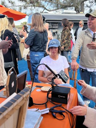 Woman wearing a Vineyard cap seated at a beachside pop-up market stall, surrounded by people, art displays and a vintage trailer backdrop, with an Orion machine and tools on an orange tablecloth.