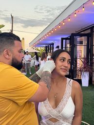 Woman in white lace dress getting her ear pierced by a gloved, tattooed piercer at an outdoor patio party under string lights and purple evening lighting.