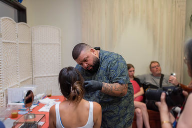 Tattooed body piercer wearing gloves carefully pierces a woman’s ear in a small indoor studio, with a mirror and piercing supplies on a table and onlookers including a photographer watching.