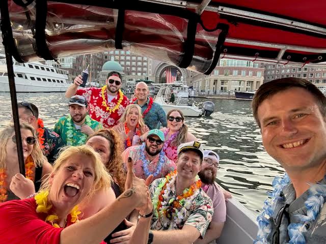 Boat party selfie: group wearing Hawaiian shirts and leis smiling aboard a harbor cruise with yachts and waterfront buildings in the background.