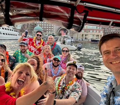 Boat party selfie: group wearing Hawaiian shirts and leis smiling aboard a harbor cruise with yachts and waterfront buildings in the background.