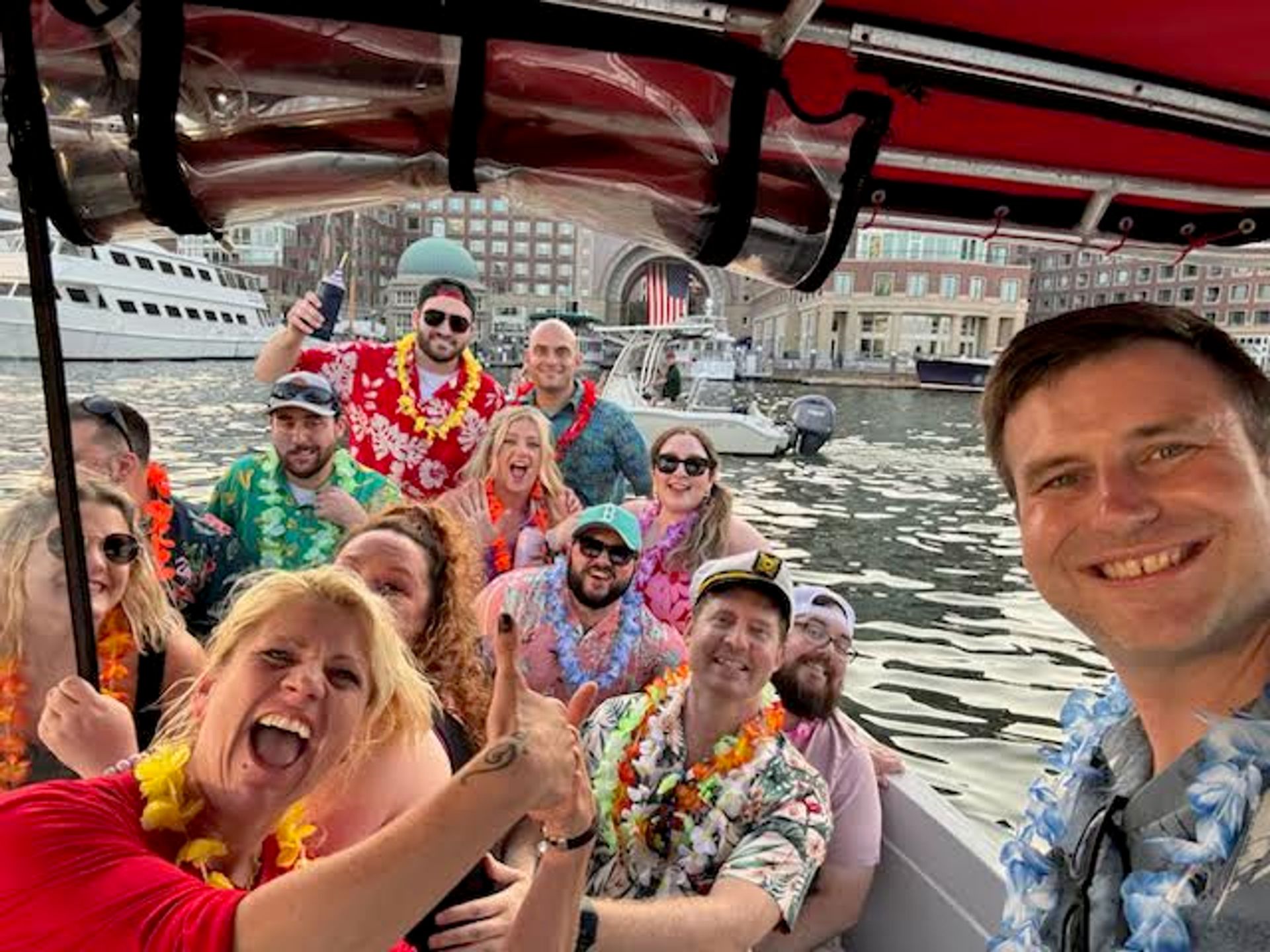 Boat party selfie: group wearing Hawaiian shirts and leis smiling aboard a harbor cruise with yachts and waterfront buildings in the background.