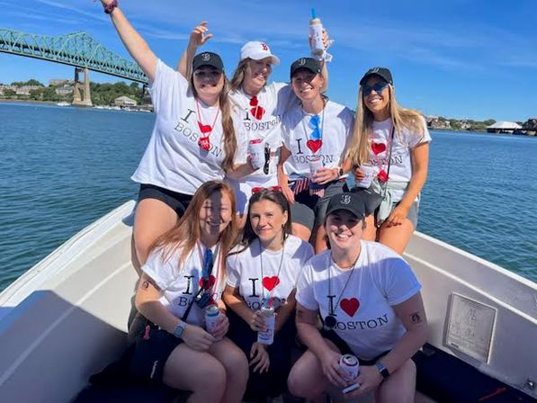 Seven friends in matching “I ♥ Boston” T‑shirts enjoying a sunny boat ride in Boston Harbor, holding drinks and posing with a green steel bridge in the background.