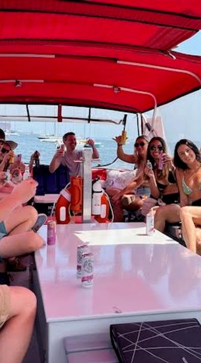 Group of friends relaxing and raising drinks on a small motorboat with a red canopy in a sunny harbor, sailboats and coastline visible in the background.