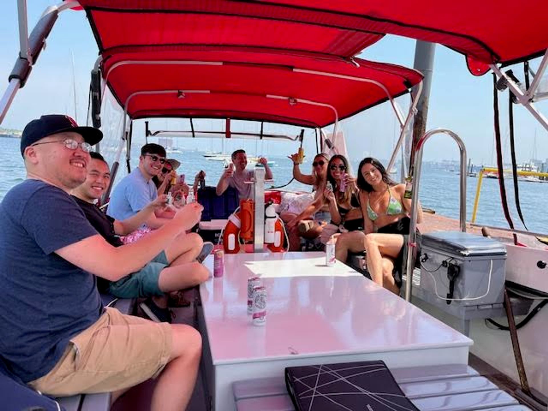Group of friends relaxing and raising drinks on a small motorboat with a red canopy in a sunny harbor, sailboats and coastline visible in the background.
