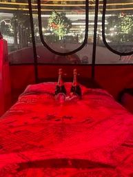 Romantic red-lit enclosed pod with a bed draped in a bright red blanket, two champagne bottles and glasses on the pillows, and round windows framing twinkling outdoor lights