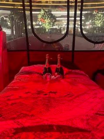 Romantic red-lit enclosed pod with a bed draped in a bright red blanket, two champagne bottles and glasses on the pillows, and round windows framing twinkling outdoor lights