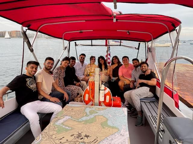 Group of friends smiling aboard a small red‑canopied tour boat over a nautical chart with life rings, cruising past a harbor with sailboats and waterfront buildings.