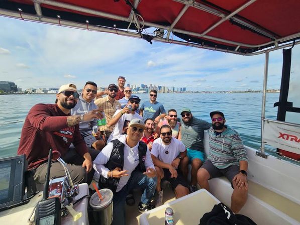 Smiling group of friends on a motorboat in calm harbor waters, holding drinks and posing with a distant city skyline under a sunny blue sky
