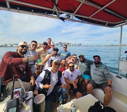 Smiling group of friends on a motorboat in calm harbor waters, holding drinks and posing with a distant city skyline under a sunny blue sky