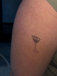 Close-up of a small minimalist martini glass tattoo on a freckled upper arm, thin linework and subtle shading with a green tank top edge visible