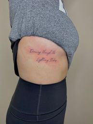 Close-up of a fresh cursive rib tattoo on a side torso reading 'Dancing through the Lightning Strikes', mild redness around the script, grey top lifted and black leggings visible.
