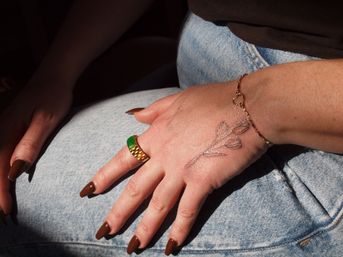 Close-up of a hand resting on light-wash denim jeans, featuring brown almond-shaped nails, a chunky gold ring with a green stone, a delicate three-bud floral tattoo on the back of the hand, and a heart-link gold bracelet.