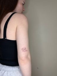 Small minimalist line-art teacup tattoo with a tiny monogram on the inner upper arm of a person wearing a black tank top against a neutral wall
