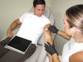 Patient reclining in a medical clinic chair receiving arm injection from a gloved clinician, tablet resting on his lap.